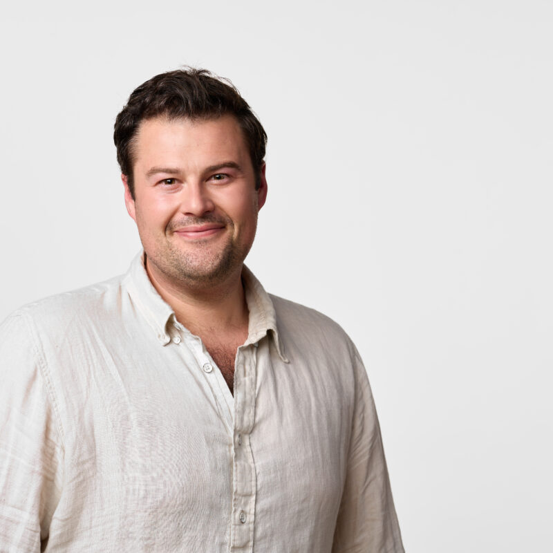 a smiling man with brown hair and stubble stands in front of a plain white wall in a white linen button down shirt