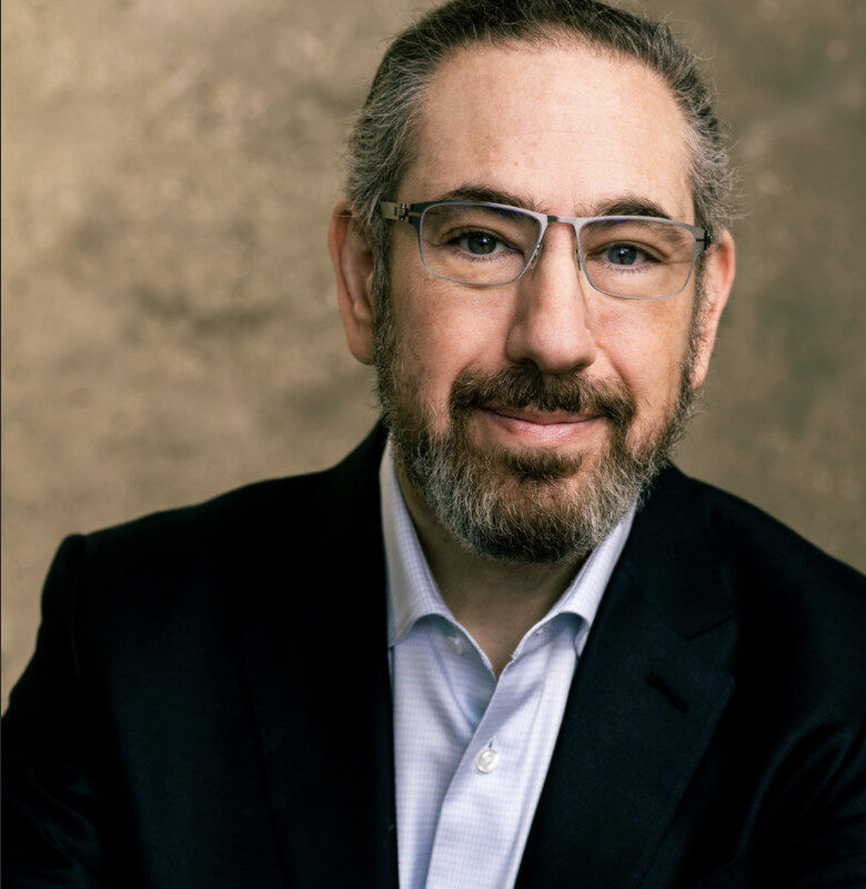 A man with glasses and a beard sits in front of a textured wall, looking thoughtfully to the side.