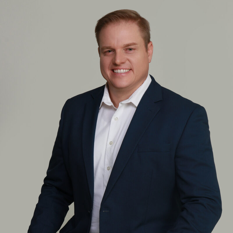 A man in a formal suit sits on a stool, looking confidently at the camera.