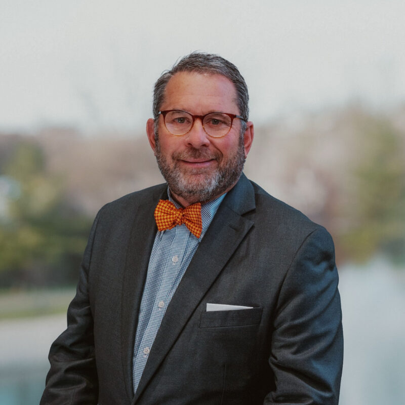 A man wearing glasses and a bow tie stands in front of a decorative fountain, smiling at the camera.