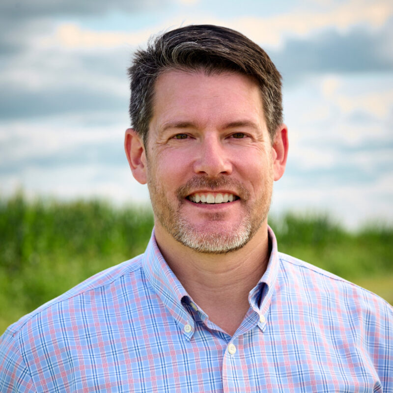 headshot of smiling Caucasian man in front of green field and blue sky with brown hair and a blue button down shirt