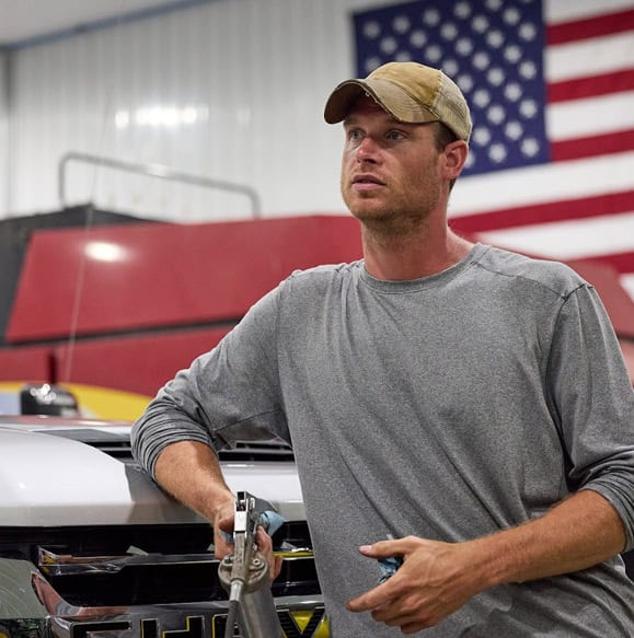 A farmer leans against a tractor with a USA flag on the wall behind him