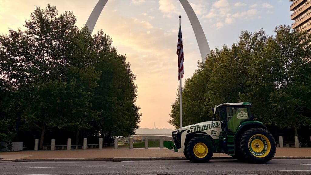 a tractor that says "Thanks" on it, in front of an American flag, which is in front of the St. Louis arch as the sun rises