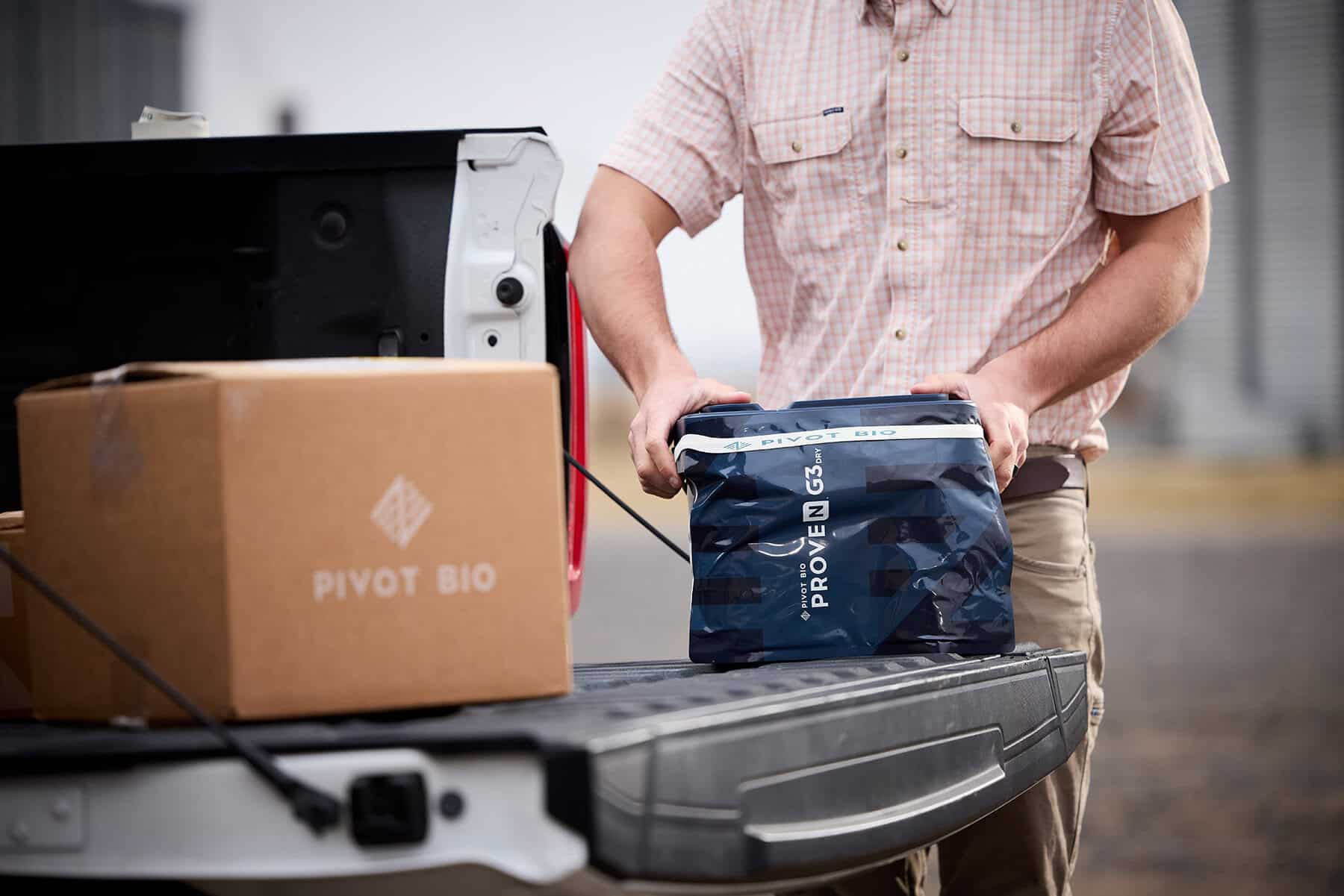 A person lifts a Pivot Bio Proven G3 bag off a pickup truck tailgate beside a box labeled Pivot Bio.