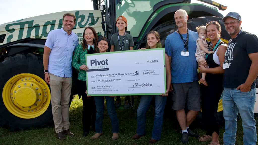 CEO Chris Abbott stands in front of the green and yellow tractor with Senator Brooke Rollins and the Fenner Family, including mom, dad, three school aged children, a toddler, and grandpa, holding a giant check made out for three thousand dollars