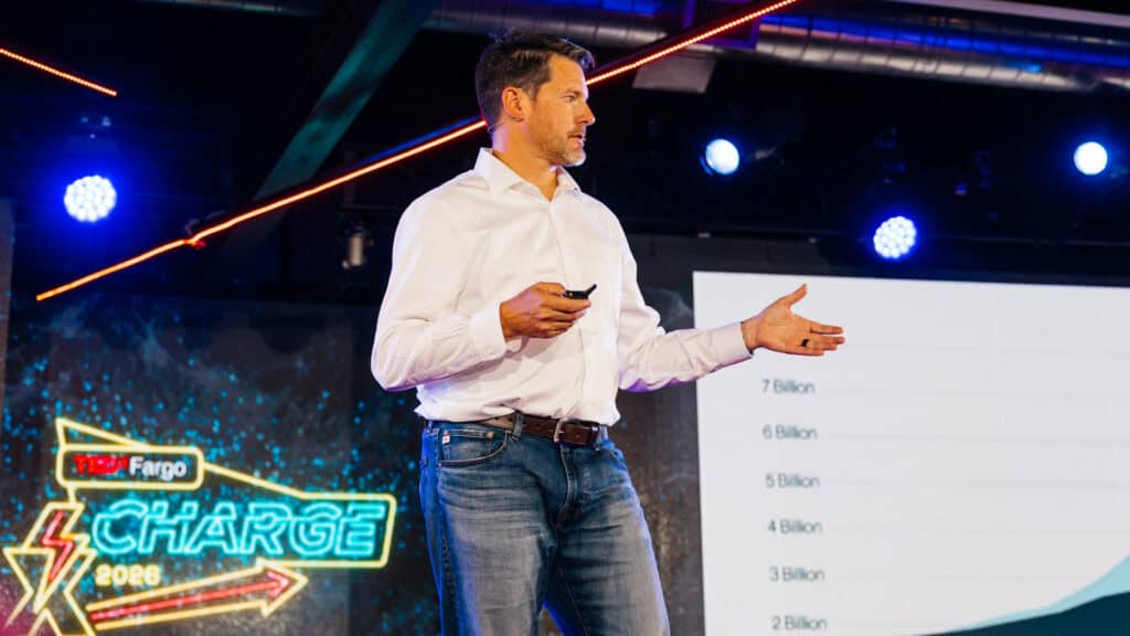 A man stands on stage, delivering a presentation to an audience, with a projector screen in the background.